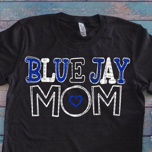 Op de afbeelding: Zwart T-shirt met de tekst "BLUE JAY MOM" in witte en blauwe letters met een blauw hart in het midden van het woord "MOM".