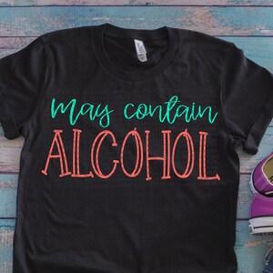 May include: Black t-shirt with the text "may contain ALCOHOL" in turquoise and red lettering. The shirt is laid flat on a blue wooden surface with a pair of purple Converse shoes and denim shorts.
