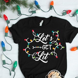 以下が含まれることがあります： カラフルなクリスマスライトに囲まれた「Let's Get Lit」という白いグラフィックがプリントされた黒いTシャツです。
