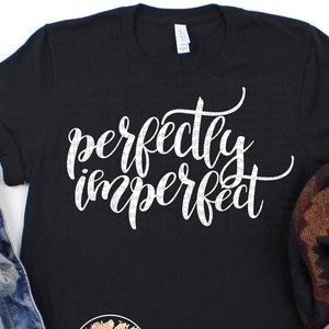 May include: Black t-shirt with the text "perfectly imperfect" in white script font. The shirt is lying flat on a white surface with a pair of brown suede ankle boots and a denim jacket.