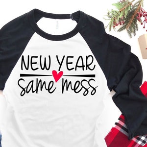 以下が含まれることがあります： 白と黒のベースボールTシャツに「新年、同じ混乱」の文字と赤いハートが描かれています。Tシャツは赤と黒のチェック柄の毛布の上にあり、青いジーンズと赤いギフトボックスが置かれています。