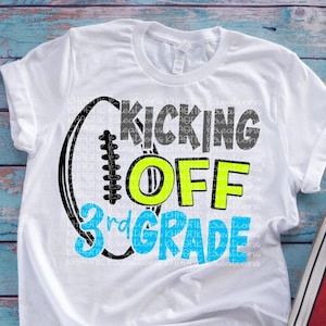 Puede incluir: Camiseta blanca con un gráfico de fútbol negro y blanco y el texto "Kicking Off 3rd Grade" en azul, amarillo y gris. La camiseta está sobre una superficie de madera azul y blanca con un par de zapatos Converse rojos.