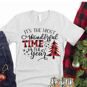 May include: A gray t-shirt with the text "It's the most wonderful time of the year" in black with a red and black plaid Christmas tree graphic. The text "Khaotik Kreations" is printed in black on the bottom right corner of the shirt.