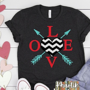 May include: A dark grey t-shirt with a red, black, and white graphic design of the word "LOVE" with a chevron patterned heart and turquoise arrows. The shirt is displayed on a white surface with a pair of white sneakers and blue jeans.
