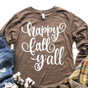 Może przedstawiać: Brązowa koszulka z długim rękawem z napisem "happy fall y'all" w białych literach. Koszulka leży na białym tle z dżinsami, pledową narzutą i kulkami bawełny.