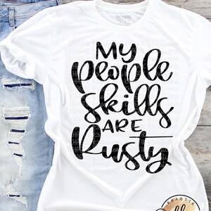 May include: White t-shirt with black text that reads "My people skills are rusty". The shirt is laying on a wooden surface with a pair of blue jeans and a small logo in the bottom right corner.