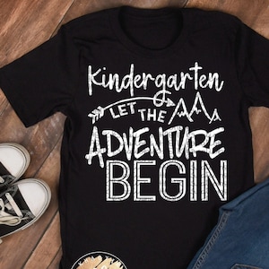 May include: Black t-shirt with white text that reads "Kindergarten Let the Adventure Begin" with a small arrow and mountain graphic.