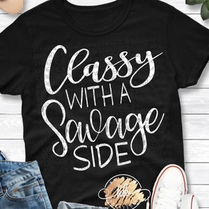 May include: Black t-shirt with the white text "Classy With A Savage Side". The shirt is paired with blue jeans, white sneakers, and sunglasses. A succulent plant is visible in the background.
