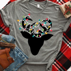 May include: A gray t-shirt with a black silhouette of a reindeer with colorful Christmas lights wrapped around its antlers. The text "Khaotik Kreations" is printed on the bottom of the shirt.