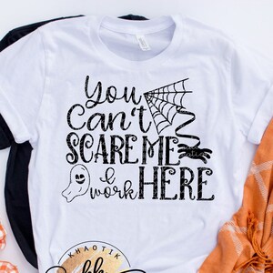 May include: White t-shirt with black text that reads "You Can't Scare Me I Work Here" with a ghost and spider graphic. The shirt is on a white surface with a black and white checkered fabric and an orange and white checkered fabric.