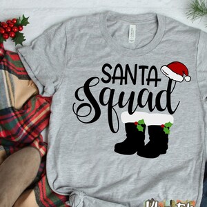 Puede incluir: Camiseta gris con el texto "Santa Squad" en letra negra. El texto está decorado con un gorro de Papá Noel rojo, ribete de piel blanca y acebo verde. Dos botas negras con ribete de piel blanca y acebo verde están debajo del texto.