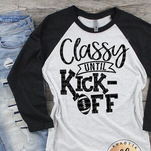 May include: A white and black baseball-themed t-shirt with the text "Classy Until Kick-Off". The shirt has a black and white color scheme and features a football graphic. The shirt is a raglan style with black sleeves and a white body.