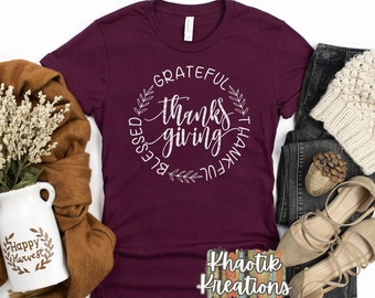 Puede incluir: Camiseta color burdeos con un gráfico circular blanco que dice "Grateful, Thankful, Blessed" con una corona de hojas alrededor del texto. La camiseta está sobre una superficie blanca con una bufanda marrón, un par de zapatos marrones, unos vaqueros y un gorro blanco de punto.
