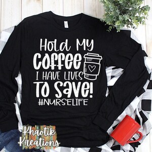 Puede incluir: Camiseta negra de manga larga con texto blanco que dice "Hold My Coffee I Have Lives To Save! #NURSELIFE". Una taza de café con un corazón está representada junto al texto. La camiseta está sobre una superficie a cuadros blancos y negros.