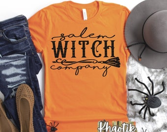 Può includere: Una maglietta arancione con la scritta "Salem Witch Company" in corsivo nero, abbinata a jeans blu strappati, ballerine beige, un cappello grigio e decorazioni di Halloween, tra cui ragni e ragnatele. Un tema di Halloween divertente e festivo.