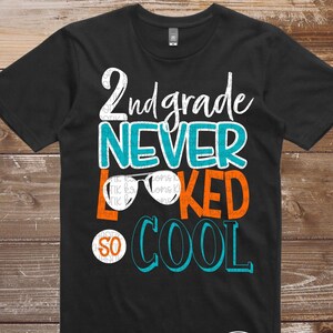 Op de afbeelding: Zwart T-shirt met een grafisch ontwerp met de tekst "2nd grade never looked so cool" in oranje, turquoise en witte tekst. De grafiek bevat een zonnebril.