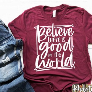Puede incluir: Una camiseta de color granate con el texto "Believe there is good in the world" impreso en blanco. La camiseta est&aacute; doblada y colocada sobre una superficie de madera blanca.