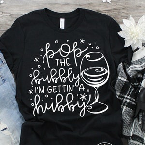 Può includere: Maglietta nera con testo bianco che recita "Pop the bubbly I'm gettin' a hibby" e un contorno bianco di un bicchiere di vino con bolle.