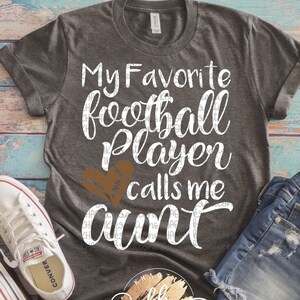 Può includere: Una maglietta grigia con il testo "My Favorite Football Player Calls Me Aunt" in lettere bianche. Un grafico a forma di cuore di football marrone è sopra la parola "aunt".