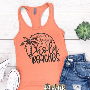 Op de afbeelding: Een koraaloranje tanktop met een zwarte afbeelding van een palmboom en zon met de tekst "Hola Beaches". De tanktop ligt op een wit houten oppervlak met een paar witte sneakers, een blauwe denim short en een kleine pot met een vetplant.