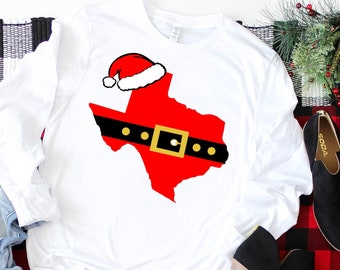May include: White long-sleeve shirt featuring a red Texas outline with a Santa hat and belt design. The belt is black with a gold buckle. The shirt has the text "Khaotik Kreations" at the bottom.