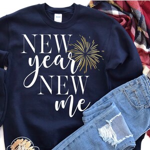 May include: Navy blue sweatshirt with the text "NEW year NEW me" in white lettering. A gold firework graphic is next to the text. The sweatshirt is laying on a white surface with a pair of blue jeans and a red and white plaid scarf.