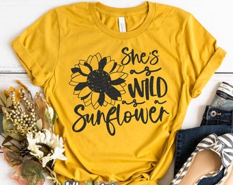 May include: A mustard yellow t-shirt with a black sunflower graphic and the text "She's as wild as a sunflower". The shirt is laid flat on a white wooden surface with a pair of blue jeans and white and black striped flats.