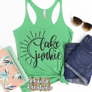 Pode incluir: Um top verde sem mangas com um gráfico preto que diz "lake junkie" com um gráfico de sol atrás do texto. O top está amarrado em um nó na parte inferior. O top está sobre um fundo branco com um par de shorts jeans azuis, um par de óculos de sol e uma embreagem rosa e verde.