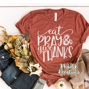 May include: A rust-colored t-shirt with the text "eat pray & give thanks" in white. The shirt is laid flat on a white wooden surface with fall-themed decor.
