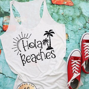 May include: White tank top with a black graphic that says "Hola Beaches" with a palm tree and sun graphic. The tank top is on a blue and red brick background.