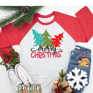 May include: A white long-sleeved t-shirt with a red and white raglan sleeve design. The shirt features a graphic of three stylized Christmas trees in green, blue, and red with white polka dots. The text "merry CHRISTMAS" is written in red and black below the trees.
