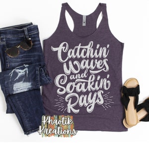 May include: Purple tank top with white text that reads "Catchin' Waves and Soak'n Rays" with a white starburst design below the text. The text is in a handwritten font. The tank top is on a white background with a pair of blue jeans and sandals.