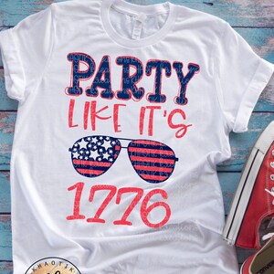 May include: White t-shirt with the text "Party Like It's 1776" in red and blue. The text is surrounded by a pair of sunglasses with an American flag design.
