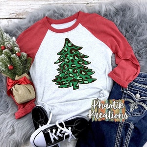May include: A white and red baseball tee shirt with a green and brown leopard print Christmas tree design. The shirt is laid out on a fuzzy gray surface with a pair of black sneakers, a small potted Christmas tree, and blue jeans with silver embellishments.