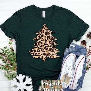 Puede incluir: Una camiseta verde oscuro con un dise&ntilde;o de &aacute;rbol de Navidad de estampado de leopardo. El texto "Khaotik Kreations" est&aacute; impreso en la parte inferior de la camiseta.