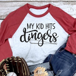 May include: A white and red baseball t-shirt with the text "My Kid Hits Dingers" in black. A baseball bat is pictured next to the text. A brown baseball glove with a white baseball is on a blue denim background.