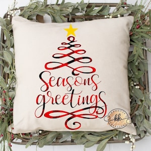 May include: A square, cream-colored pillow with a Christmas tree design. The tree is red and black plaid with a yellow star on top. The words "Seasons greetings" are written in red and black script. The pillow is surrounded by greenery.