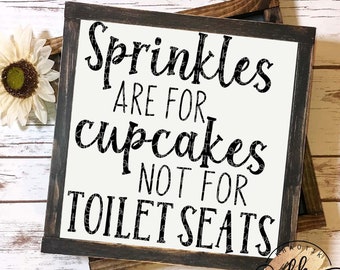 May include: A framed wooden sign with the text "Sprinkles ARE FOR cupcakes NOT FOR TOILET SEATS" in black lettering on a white background. The frame is dark brown. A decorative sunflower is in the upper left corner.