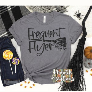 以下が含まれることがあります： 「Frequent Flyer」というテキストと、星がついたほうきの黒いシルエットがプリントされたグレーのTシャツ。右下に「Khaotik Kreations」というテキストがあります。