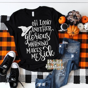 May include: Black t-shirt with white text that reads "Oh look! Another glorious morning makes me sick". The text is in a distressed font and includes a witch's hat graphic. The shirt is laid out on a black and white checkered surface with a pair of black converse shoes and blue jeans with distressed details.