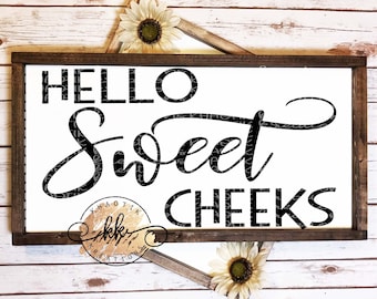 May include: A wooden sign with the text "Hello Sweet Cheeks" in black script font. The sign is on a white wood background with sunflowers.
