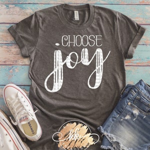 May include: A gray t-shirt with the text "Choose Joy" printed in white. The word "Joy" is stylized with a white outline and filled with a pattern of small white lines. The t-shirt is paired with denim shorts and white sneakers.