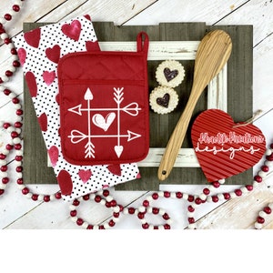May include: Red and white kitchen pot holder with an arrow and heart design. The pot holder is on a white and black polka dot kitchen towel with red hearts. A wooden spoon, red heart-shaped sign with the text "Khaotik Kreations Designs", and two cookies are also in the image.