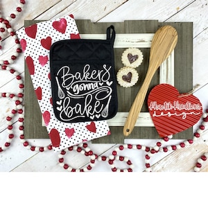 May include: A black oven mitt with white text that reads "Bakers gonna bake" with a heart design. The oven mitt is on a white and red polka dot kitchen towel with red hearts. There is a wooden spoon, two cookies, and a red heart-shaped sign that reads "Khaotik Kreations Design".
