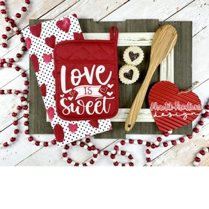 May include: Red and white kitchen towel with a heart pattern and a red pot holder with the text "Love is Sweet" in white. The pot holder has a white ribbon with a heart on each end. There is a wooden spoon, a red heart-shaped ornament, and two cookies with red hearts on top.
