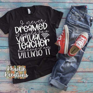 May include: Black t-shirt with white text that reads "I never dreamed I would be a virtual teacher but here I am killing it". The shirt is laying on a wooden surface with a pair of blue jeans and red converse shoes.