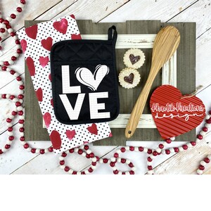 May include: A black oven mitt with the word "LOVE" in white, a red heart-shaped decoration, a wooden spoon, and a polka dot and heart-patterned kitchen towel. Two heart-shaped cookies and a beaded garland complete the Valentine's Day theme.