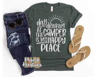 May include: A green t-shirt with white text that reads "day drinking at the camper is my happy place". The text is in a distressed font and includes a sun graphic. The shirt is laying on a white surface with a pair of brown sandals and blue jeans.