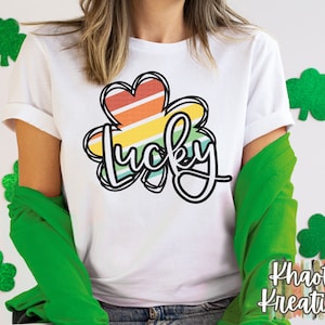 以下が含まれることがあります： 虹色のストライプのクローバーと黒い文字で「lucky」と書かれた白いTシャツ