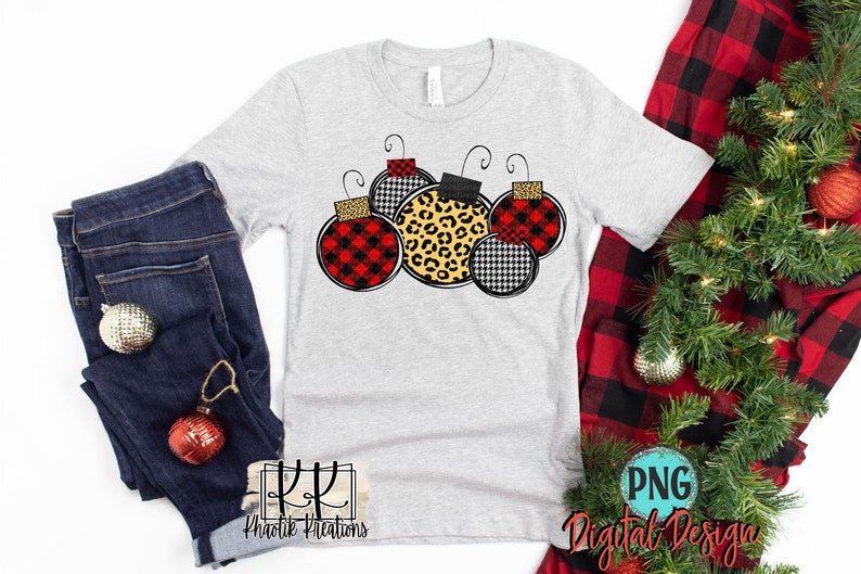May include: A gray t-shirt with a design of five Christmas ornaments. The ornaments are red and black plaid, black and white houndstooth, and leopard print.